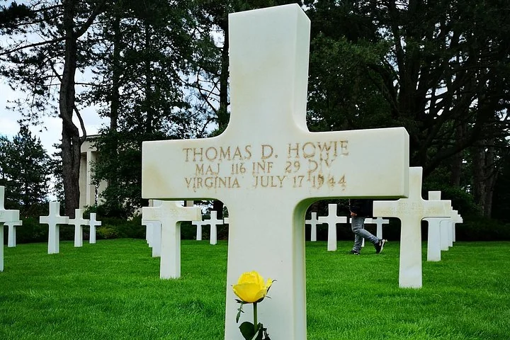 A white marble cross engraved Thomas D. Howie, Maj 116 Inf 29 Div, Virginia, July 17 1944, with a yellow rose at its foot