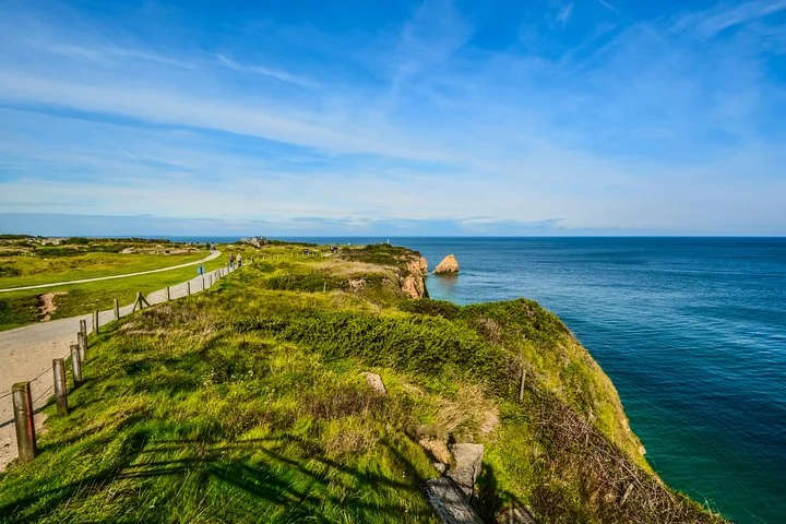 A grassy clifftop path with a wooden fence above deep blue sea, a rock stack off the point