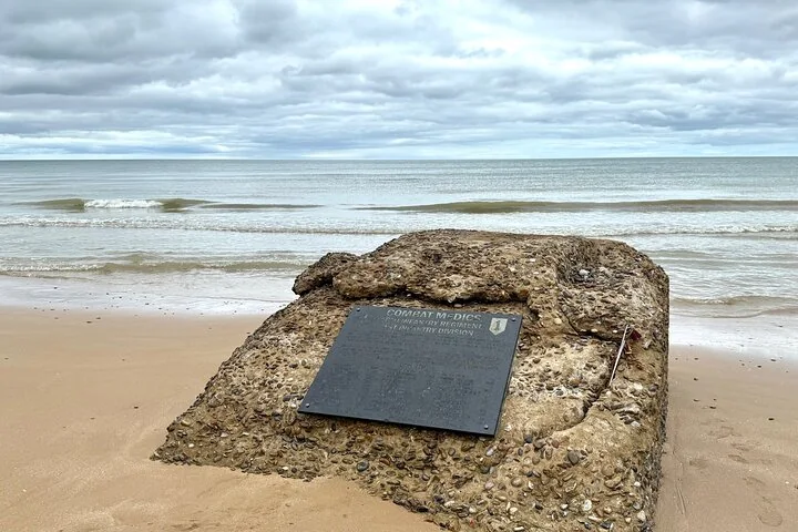 A memorial plaque to the combat medics of the 1st Infantry Division set on a concrete block on the sand, with the sea behind