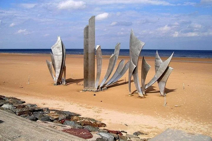 A tall stainless steel sculpture of rising blades standing on a sandy beach