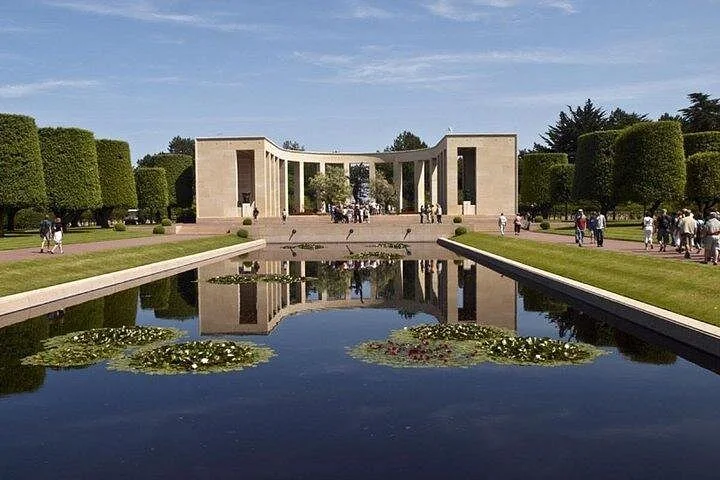 A stone memorial colonnade reflected in a long pool with water lilies, lined by clipped trees