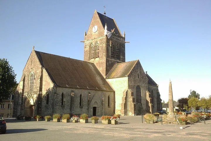 A stone church with a square tower seen across a village square, a parachute on the steeple