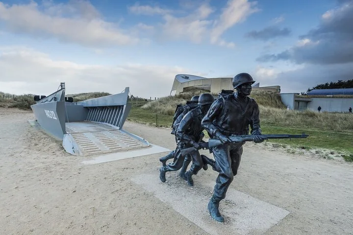 Bronze statues of soldiers running up from a landing craft, with a museum building in the dunes behind